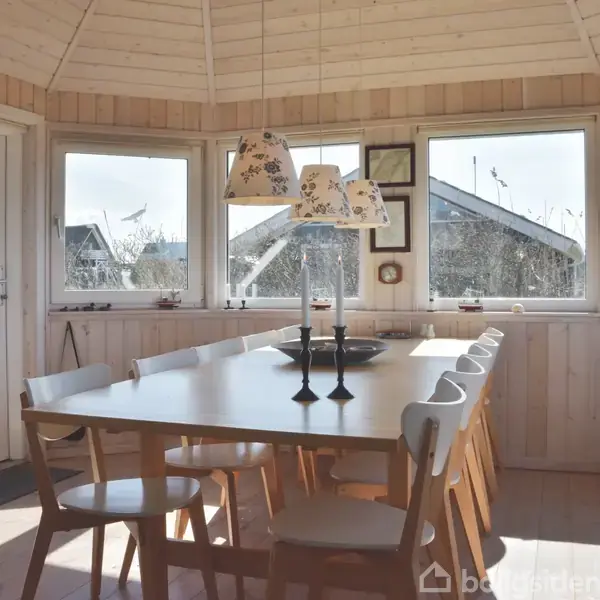 Et lyst træbord omgivet af otte stole står i et solbeskinnet rum med trævægge. Store vinduer giver udsigt til huse udenfor. En lampe hænger over bordet.