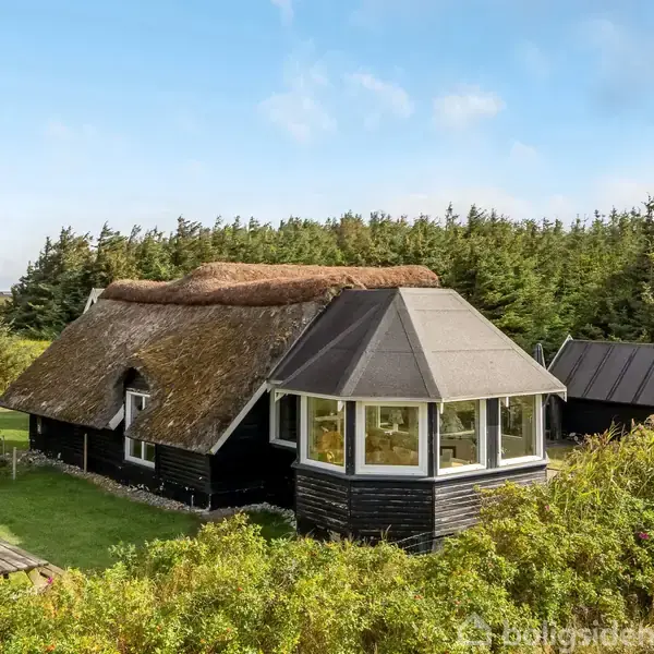 Et stråtækt hus med stort vinduesparti står stille omgivet af tætte grønne buske og træer. En flagstang og et picnicbord ses i forgrunden under en blå himmel.