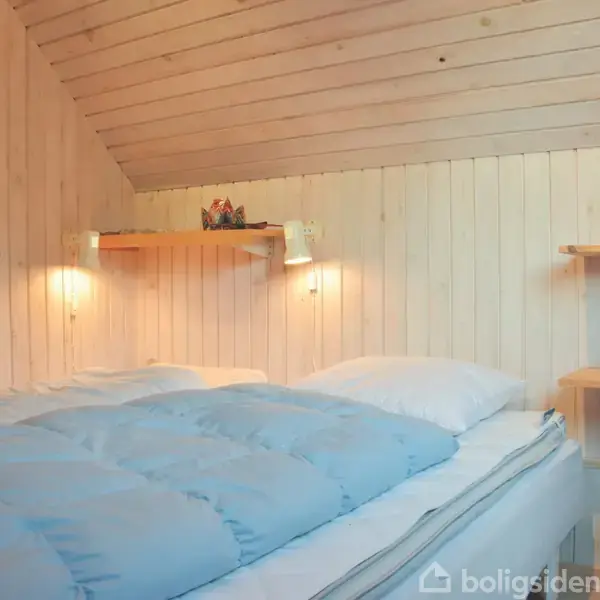 En seng med lyseblåt sengetøj står i et træbeklædt soveværelse. To væglamper lyser op omkring en hylde over sengen, og der hænger en farverig plakat på væggen.