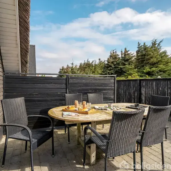 Rundt havebord er omkranset af flettede stole. Bordet er dækket med morgenmad i solrig terrasse, omkranset af sorte træhegn, tæt ved mørke husfacader og grønne træer i baggrunden.
