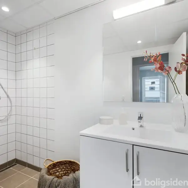 Håndvask med spejl reflekterer en lys og moderne badeværelseindretning. Ved siden af er bruseområdet med hvide fliser. En kurv og en vase med blomster står på vasken.