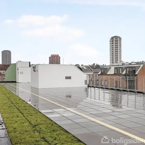 Tagterrasse med grønne planter langs siden og våde fliser i midten, omkranset af et metalkrækkelværk. Baggrund viser bybygninger og højhuse under en overskyet himmel.