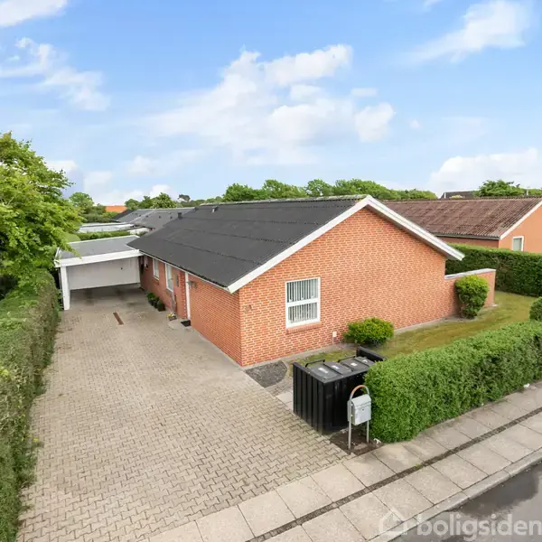 Rødt murstenshus med sort tag står stille i et forstadskvarter, omgivet af velplejet hæk og brolagt indkørsel. Bil opbevaret under carport til venstre. Skyfri himmel over området.