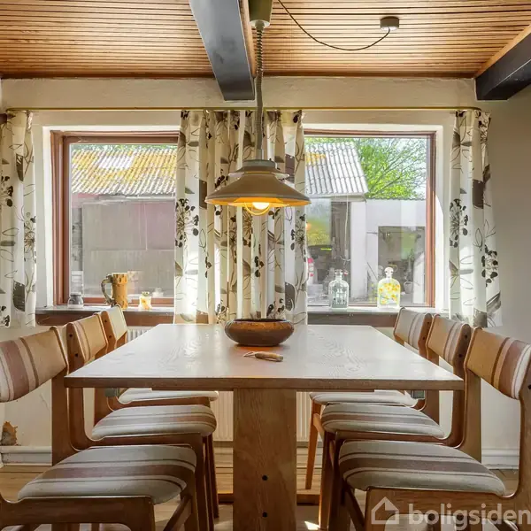 Et træbord er omgivet af otte stole, placeret i en spisestue med store vinduer og udsigt til en have. En loftslampe hænger over bordet, og en reol er til højre.