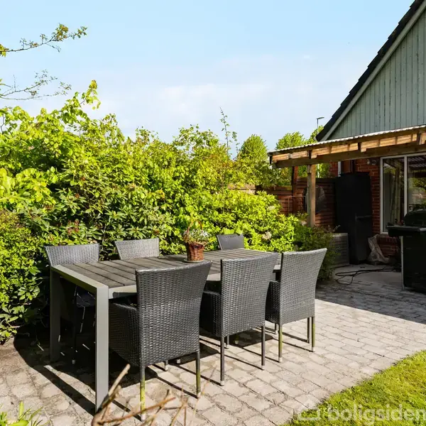 Terrasse med sort bord og seks stole, står på flisebelagt område omkranset af grønne buske. Huset er delvist synligt med røde mursten og et vindue i baggrunden.
