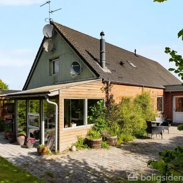 Et to-etagers hus med en rødt tegltag og vinduer. En terrasse med havemøbler i en frodig haveomgivelse. Satelitantenne og skorstene på taget.