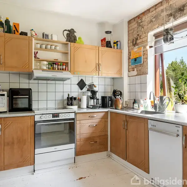 Et køkken med træskabe og hvide fliser. En ovn og en mikrobølgeovn står på bordet. Udenfor vinduet ses en grøn have. Ikke synlig tekst.