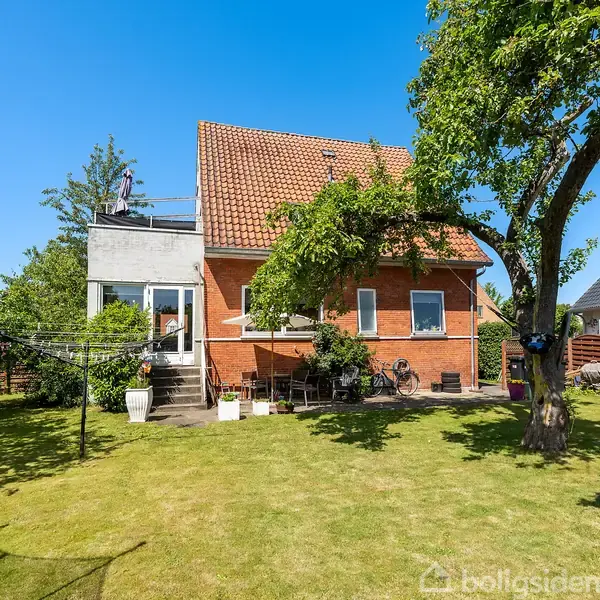 Et rødt murstenshus med en stor terrasse ses i en velholdt have. Græsplænen omkranses af træer, og solen skinner fra en klar blå himmel.