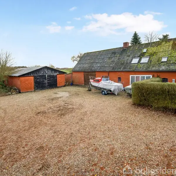 Hus med mosdækket tag og røde mure står ved en grusbelagt gårdsplads med en afdækket båd. Ved siden af husene ses en ældre, rød garage.