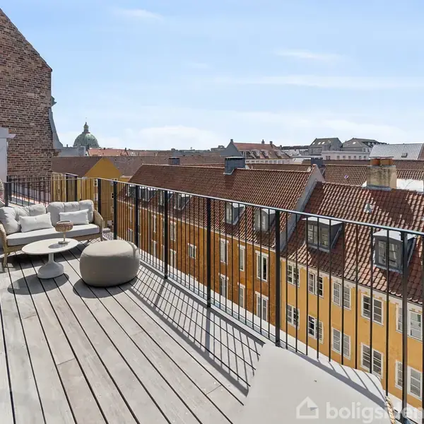 En tagterrasse med trædæk har komfortable siddemøbler og et lille bord, omgivet af gamle, murstens- og gulmalede bygninger, og med en udsigt, der inkluderer en kuppel i det fjerne.
