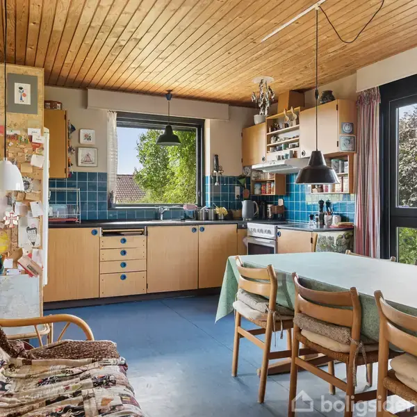 Køkkenbord med stole står i et rum med træloft og blå fliser, omgivet af skabe og komfur; grønne planter ses gennem vinduet. Billeder og noter hænger på væggen.