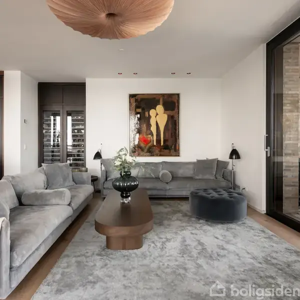 Et moderne opholdsrum med en stor grå hjørnesofa, sofabord og skammel. Kunstværk hænger på væggen. Køkkenområde i baggrunden med barstole og træelementer.