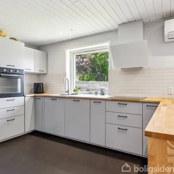 Køkken med hvide skabe og indbygget ovn. En håndvask under et vindue med udsigt til have. Bordplade i lyst træ. Væggen har hvide fliser. Klimaenhed på væggen.