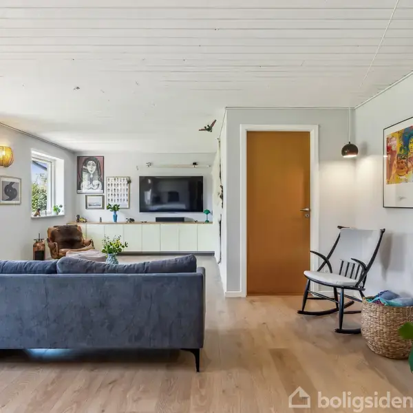Sofa står i en stue med trægulv og hvide vægge. Et TV er monteret på væggen. Rumdekorationer omfatter kunstværker, planter og en gyngestol ved siden af en kurv.