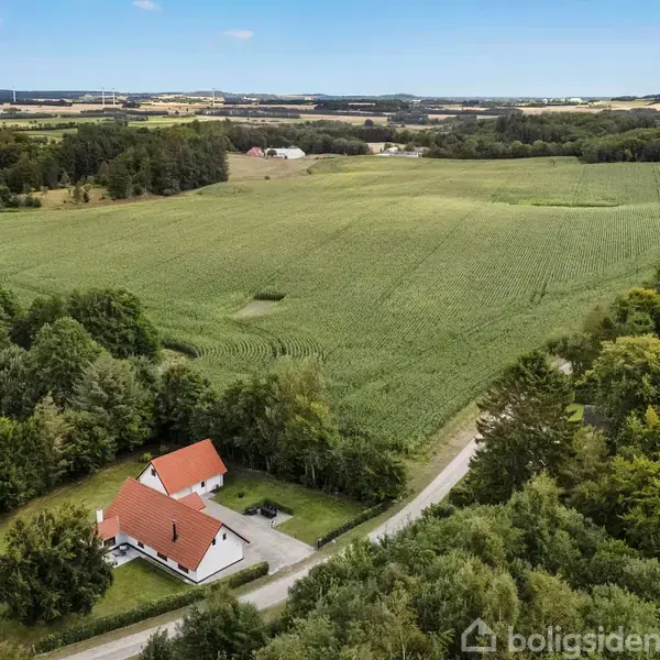 Et hvidt hus med rødt tag ligger blandt grøn vegetation ved en snoet vej. Det er omgivet af marker og skov i et landligt landskab under en blå himmel med enkelte skyer.