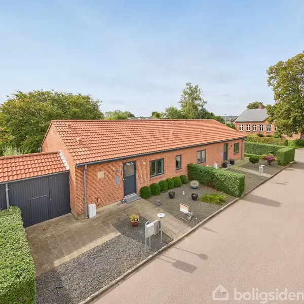Rødt murstenshus med rødt tegltag står langs en rolig villavej med fortov. Omgivet af hæk og have, med tilhørende garage på venstre side. Klar, solrig dag.