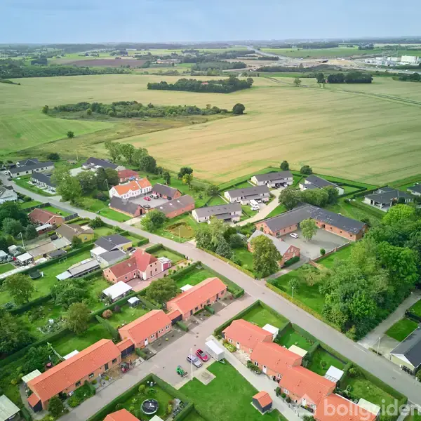 Beboelseshuse med røde tage ses fra luften, stilleliggende langs en vej. Omkringliggende grønne marker strækker sig ud i horisonten. Vegetation og små haver omgiver husene, mens marker omkranser området.