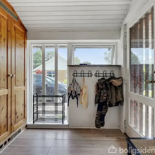 Træskab står i gangareal med bøjleophæng fyldt med tøj. Gennem vinduet ses en parkeret bil og et hus i baggrunden. Dør til højre leder ud.