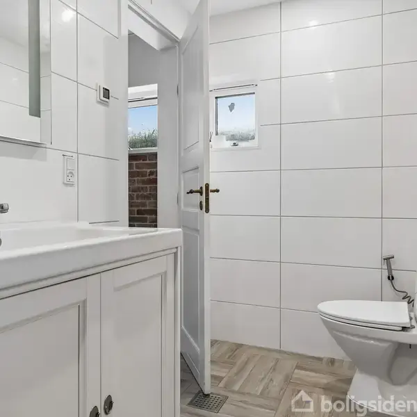 Hvidt badeværelse med en vask med skab og et toilet. Åben dør fører ud til overdækket område med murstensvæg. Området er lyst op af et lille vindue.