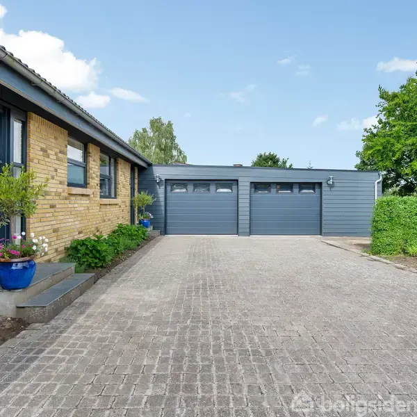 Et hus af gule mursten med to blå garageporte. Foran huset er der en brostensbelagt indkørsel, omgivet af grønne buske og planter. Himmel er klar med enkelte skyer.
