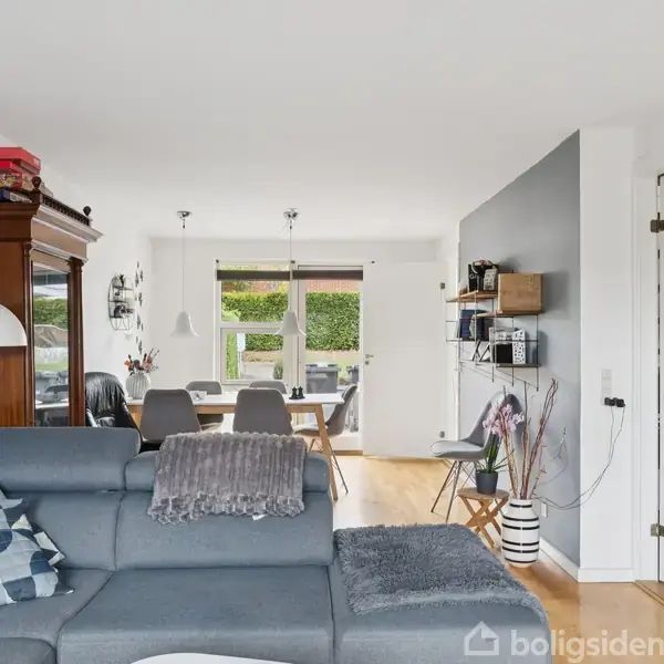 Grå sofa med puder står i stuen, hvor et spisebord med stole ses i baggrunden. Rummet er lyst med hvide vægge og vinduer, der lukker naturligt lys ind.