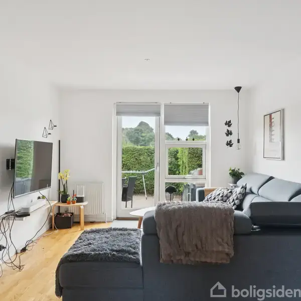 Stue med en grå hjørnesofa, hvor et tæppe og puder ligger. En fladskærms-tv er vægmonteret. Glasdøre åbner ud til en terrasse med udsigt over en grøn hæk.