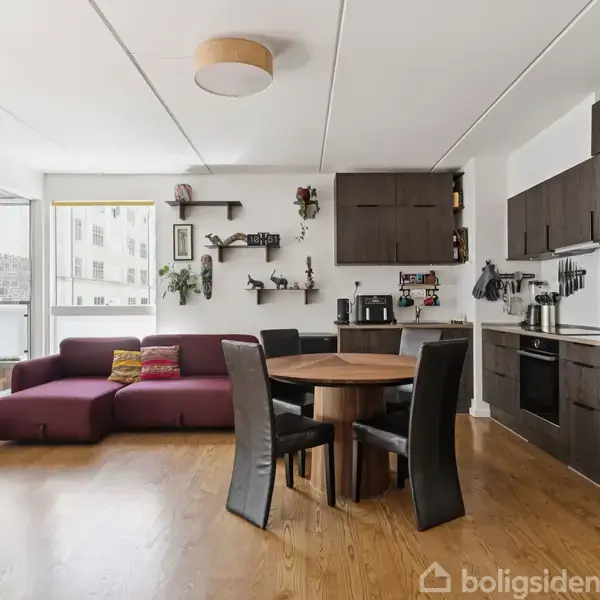 Mørkebrune køkkenmøbler i et moderne, åbent køkken-stue-område, med et rundt spisebord og fire stole. En lilla sofa står ved et stort vindue, der trækker lys ind.