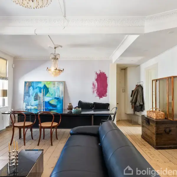 Sort lædersofa står centralt i en lys stue med trægulv, moderne kunst på væggen, og store vinduer. To træstole står ved siden af en lav bordplade med bøger og dekorationer.