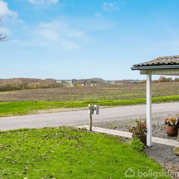 Hus med rød væg og veranda, set fra en græsplæne. Verandaen har en blomsterpotte. I baggrunden ses en vej, marker og træer under en blå himmel.