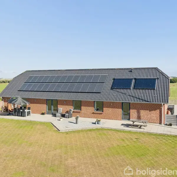 Et rødt murstenshus med solpaneler på taget står i en åben, grøn mark. En terrasse med havemøbler ses foran huset. Vejen løber langs siden af ejendommen.