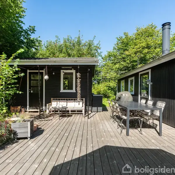 Et sort træhus står på en træterrasse, hvor et havesæt og grill er placeret. Haven er omgivet af grønne træer og buske i baggrunden.