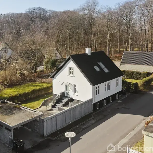 Et hvidt hus med sort tag står på en lille vej. Der er en trappe op til hoveddøren. Omkring huset er der træer og en garage til venstre.