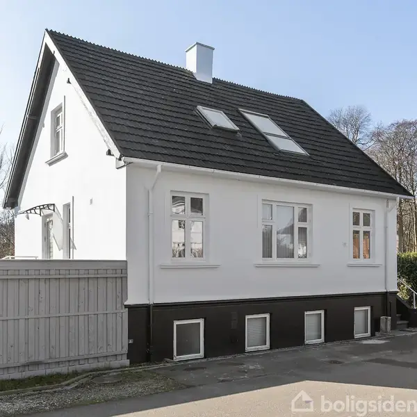 Et hvidt hus med sort tag har vinduer på taget og vægge. Det står ved siden af et træhegn og asfaltvej, omgivet af træer i baggrunden.