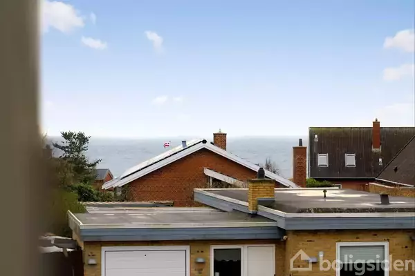 Et gult murstenshus med fladt tag står i forgrunden. I baggrunden ses et rødt hus med skråt tag, og der er udsigt til havet under en blå himmel.