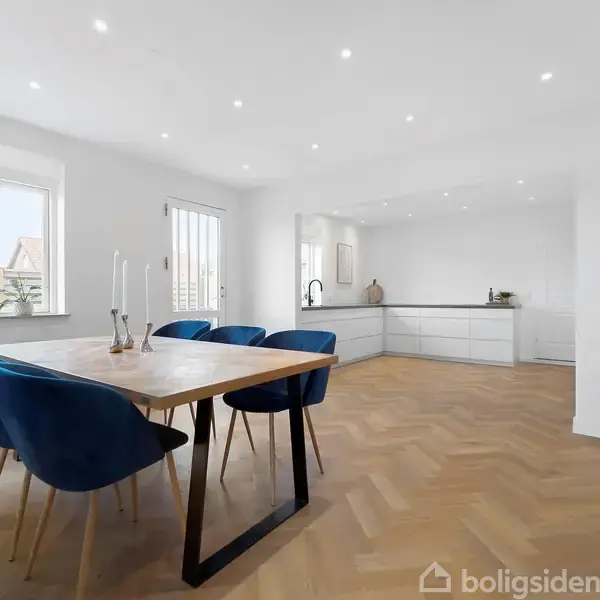 Bord med blå stole står i en spisestue. Rummet er lyst med trægulv, hvidmalede vægge, åbner til et køkken. Et hvidt ur hænger på væggen over en skænk.