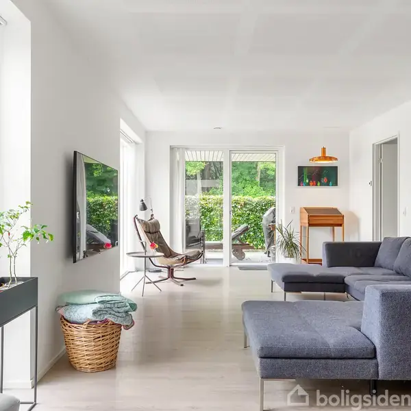Stue med grå hjørnesofa, moderne lænestol og fjernsyn på væggen. Store vinduer og glasdør åbner mod grøn have. Kurv med tæpper og planter i siderne.