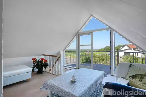 Hvidt møbleret stue med bord dækket af et blåt mønsteret dug, åbne døre til en altan, med udsigt over grønne områder og blå himmel. Blomster og sofa ses i rummet.