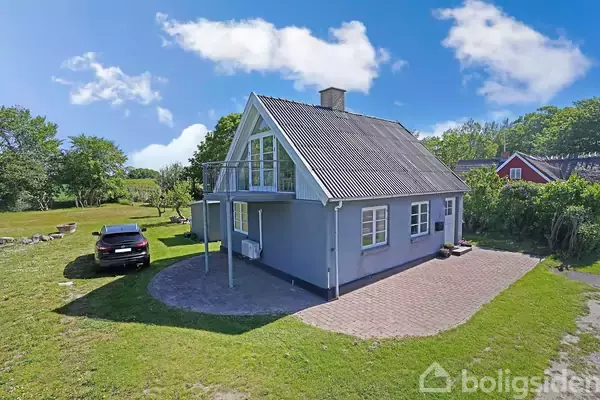Et gråt hus står på en grøn græsplæne med en sort bil parkeret ved siden af. Træer omgiver stedet, og himlen er klar med få skyer.