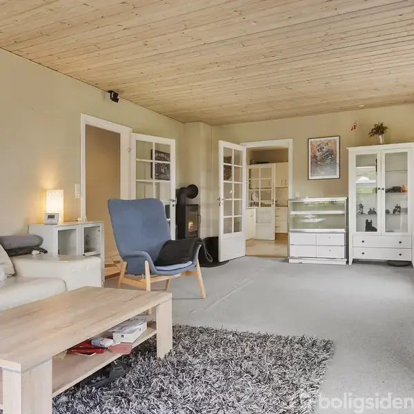 Stue med lys sofa og blå lænestol ved et træbord, omgivet af reoler og en pejs i et hyggeligt, komfortabelt rum med lyse vægge og træloft.