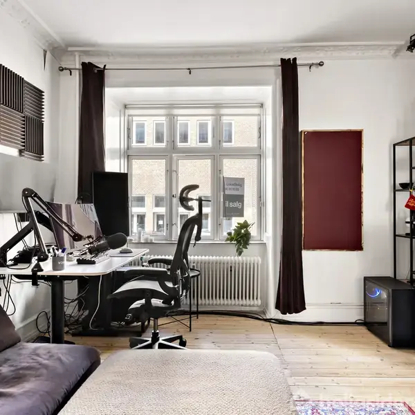 Soveværelse med en dobbeltseng dækket af et gult sengetæppe. Til venstre står en sort kommode med lampe og spejl. Et tæppe ligger på gulvet; et vindue viser bygning udenfor.
