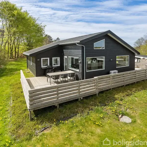 Sort sommerhus med terrasse omgiven af græsplæne og nogle træer i baggrunden. Terrasse har bord og stole, beskyttet af træhegn. Klar blå himmel ses ovenfor.