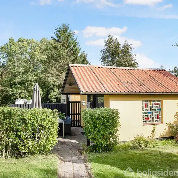 Et lille, gult hus står midt i en grøn have. En sti fører dertil, omkranset af buske. Baggrunden domineres af træer under en blå himmel.