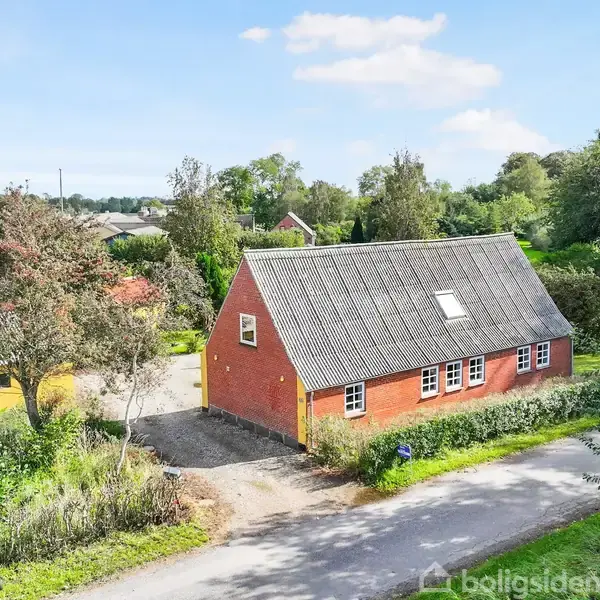 Rødt murstenshus med gråt tag står stille langs en vej omgivet af grønne træer og buske i landligt område. Der ses en gul udhusbygning i baggrunden.