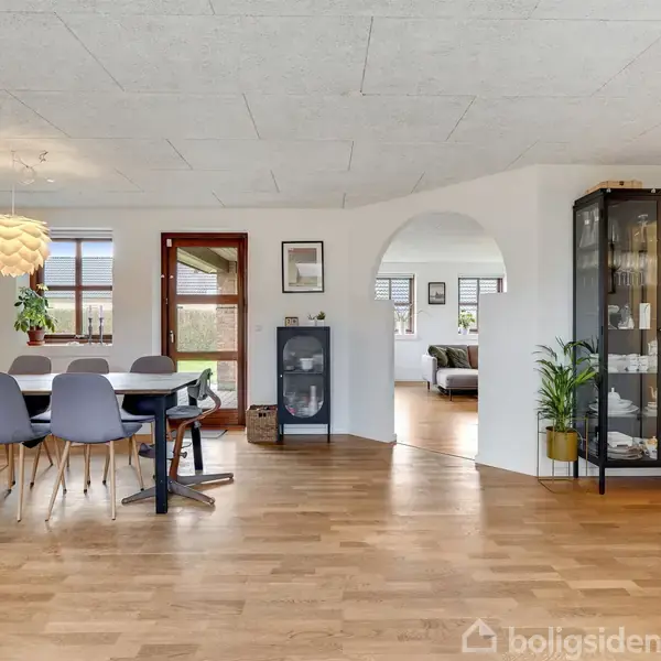Spisebord med blå stole står på trægulv i rummeligt, lyst rum. Moderne pendellampe hænger over bordet. Glasvitrine og grøn plante ses til højre. I baggrunden er en sofa.