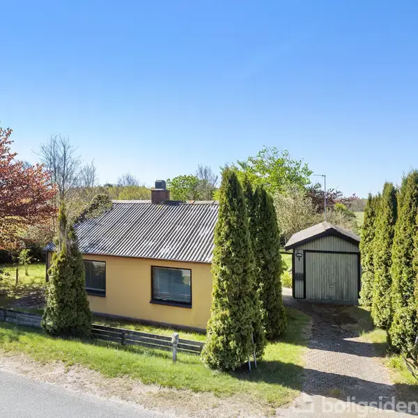 Et gult hus står omgivet af høje træer langs en stille vej. En sti fører til en lille garage i baghaven under en klar, blå himmel. Skilt med "Til salg".