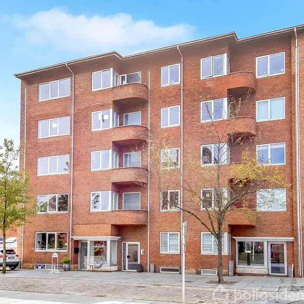 Bygning med fem etager, rød murstensfacade og små altaner, står ved en fortovsgade med få træer. Biler og cykel er parkeret foran. Klare, blå himmel i baggrunden.