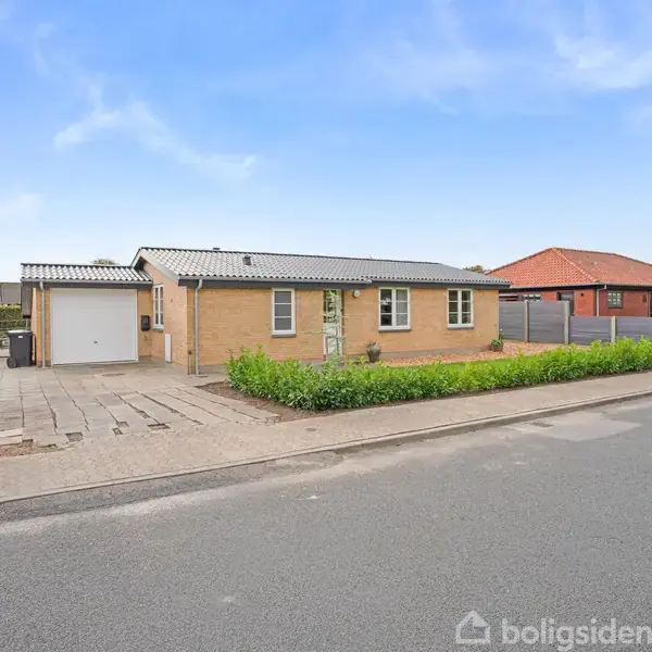 Et enkelthus i mursten står stille, omgivet af grønne buske ved fortovet i et boligområde med flere lignende huse omkring.