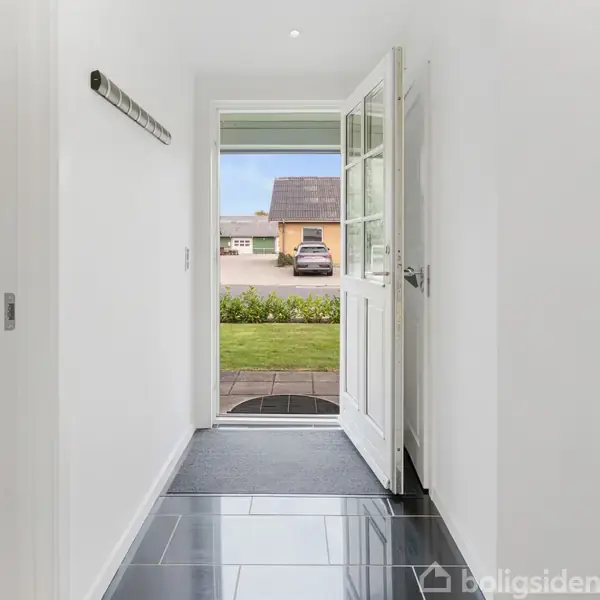 Dør står åben i en lys gang med sort flisegulv, der fører ud til en forhave. Til venstre ses et vindue og en del af en sofa.