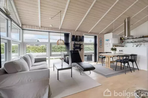 Stue med sofa og chaiselong på en lys pude, ved siden af et spisebord med stole. Store vinduer lukker lys ind. Lyse trælofter og køkken i baggrunden.