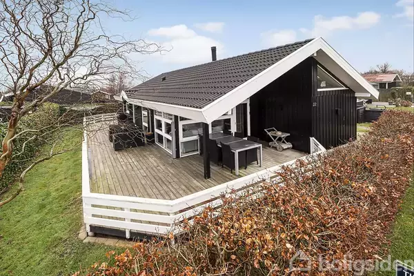 Et sort træhus med hvidt tag og terrasse omgivet af en hækkant. Huset står på en græsplæne, og himlen er let overskyet.